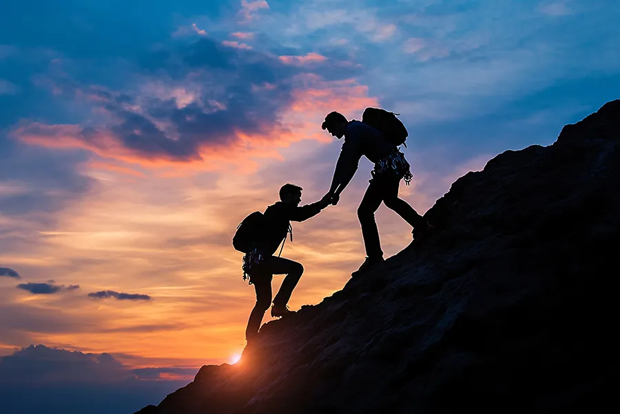 Two climbers silhouetted at sunset, with one helping the other up a rock face.