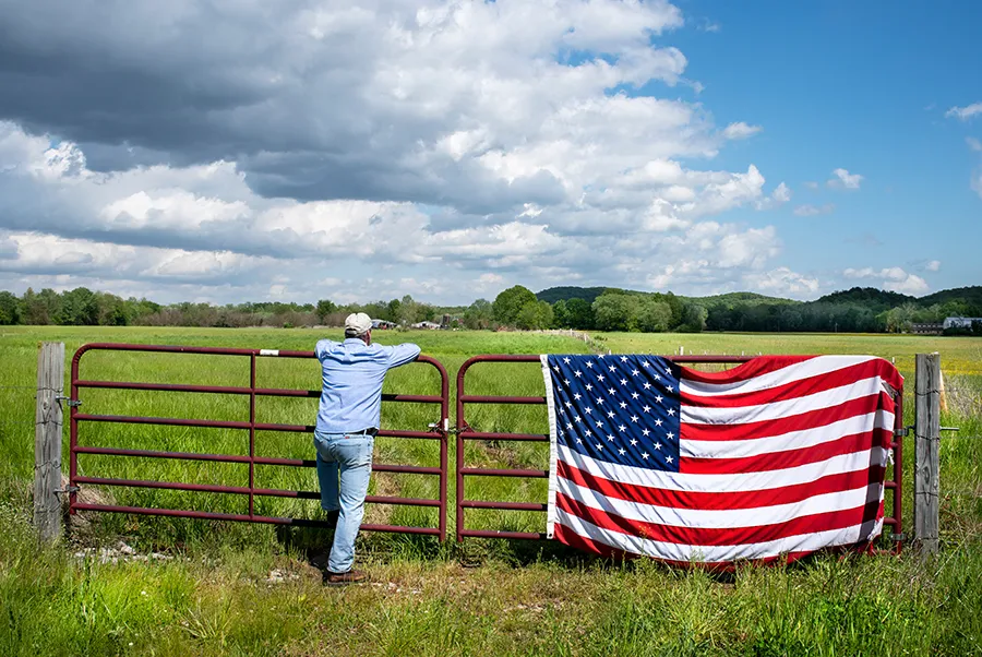 Person standing at a farm gate draping an American flag.